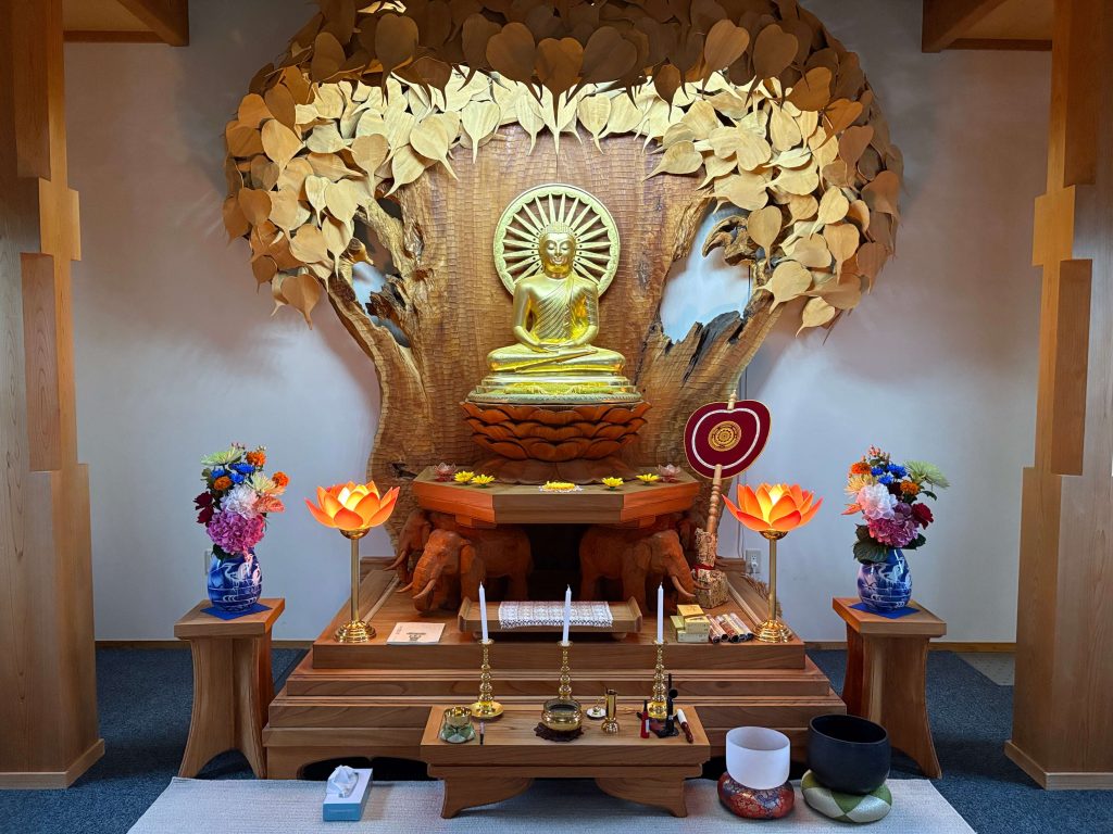 Golden Buddha statue seated in lotus position on a wooden altar, with a carved tree backdrop and gold-leaf leaves above. Vases of colorful flowers and orange lotus lamps flank the altar.