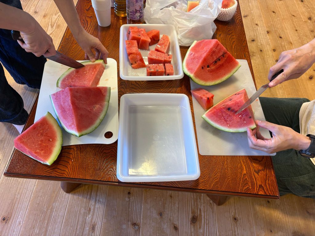 People cutting and serving watermelon slices at a wooden table; cubed watermelon in a tray and large wedges ready to eat.
