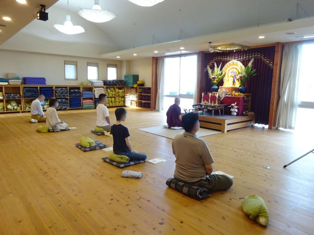 People kneeling on cushions in a bright meditation hall, facing a shrine with flowers and a statue on an altar.