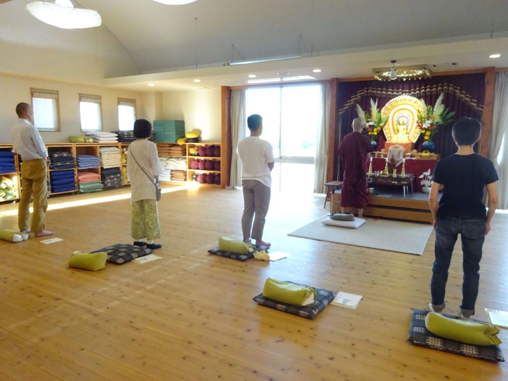 Group of people standing on cushions in a meditation hall, facing a Buddhist altar with a statue and flowers at the front.
