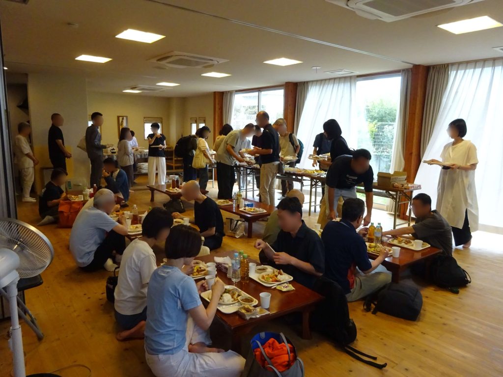 People queueing for food at a casual buffet-style gathering in a bright dining hall with windows and curtains.