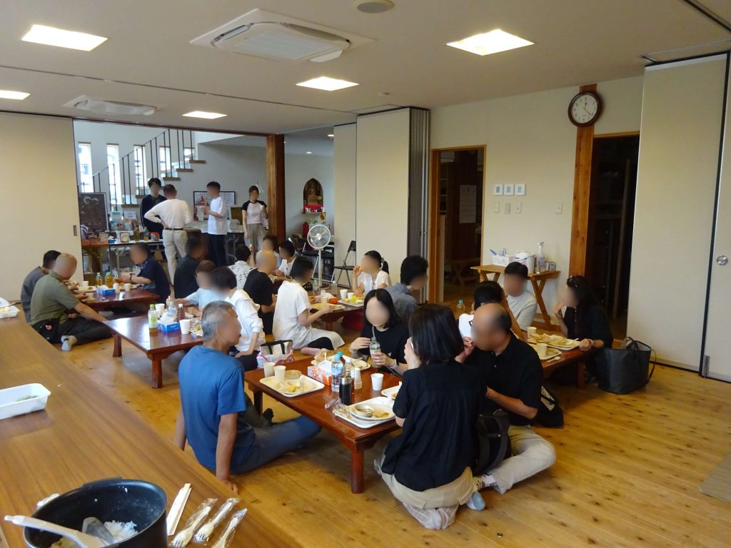 Large group of people seated on the floor and at low tables, eating a meal in a bright, open room.