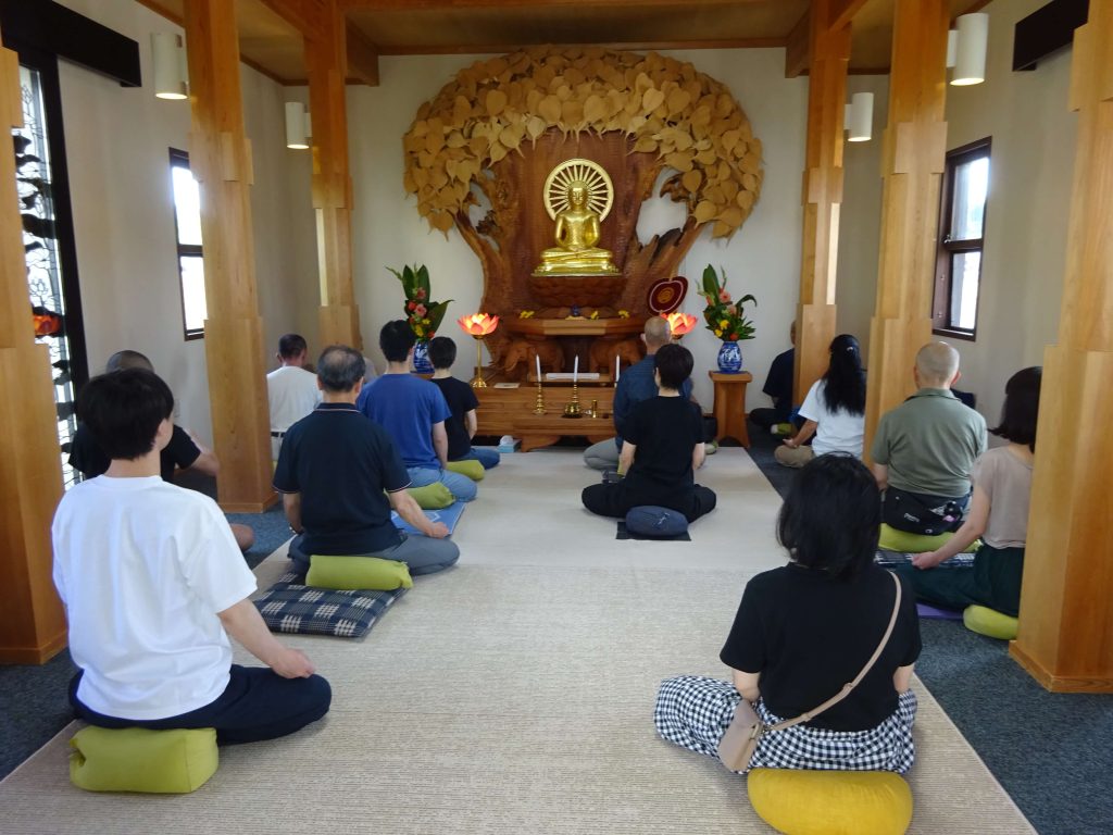 Group of people meditating on cushions facing a golden Buddha statue inside a wooden shrine room