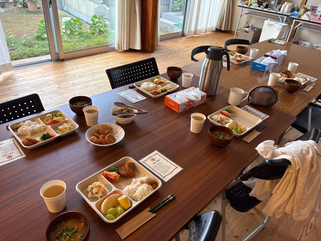 Long wooden table with multiple bento boxes, soups, fruit, and tea cups in a bright dining room by large windows.