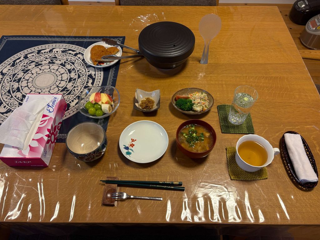 Overhead view of a wooden table with a Japanese meal: miso soup bowls, fruit bowl, fried croquettes, chopsticks, and napkins on a patterned placemat.