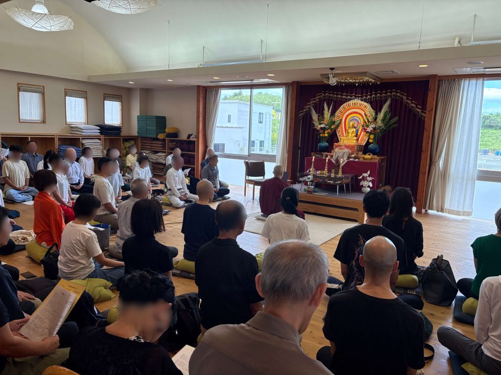 Group of people seated on cushions in a bright meditation hall, facing a Buddha altar with flowers and offerings.