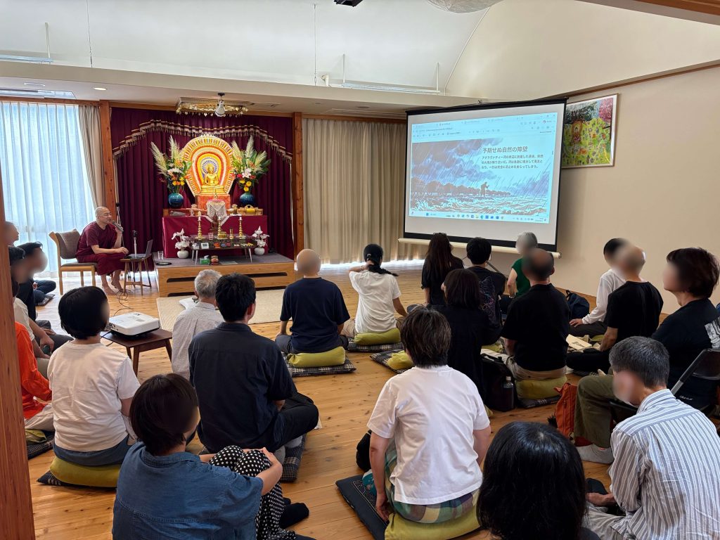 Monk in maroon robes speaks to a seated audience in a shrine room with a Buddha statue and floral altar on stage