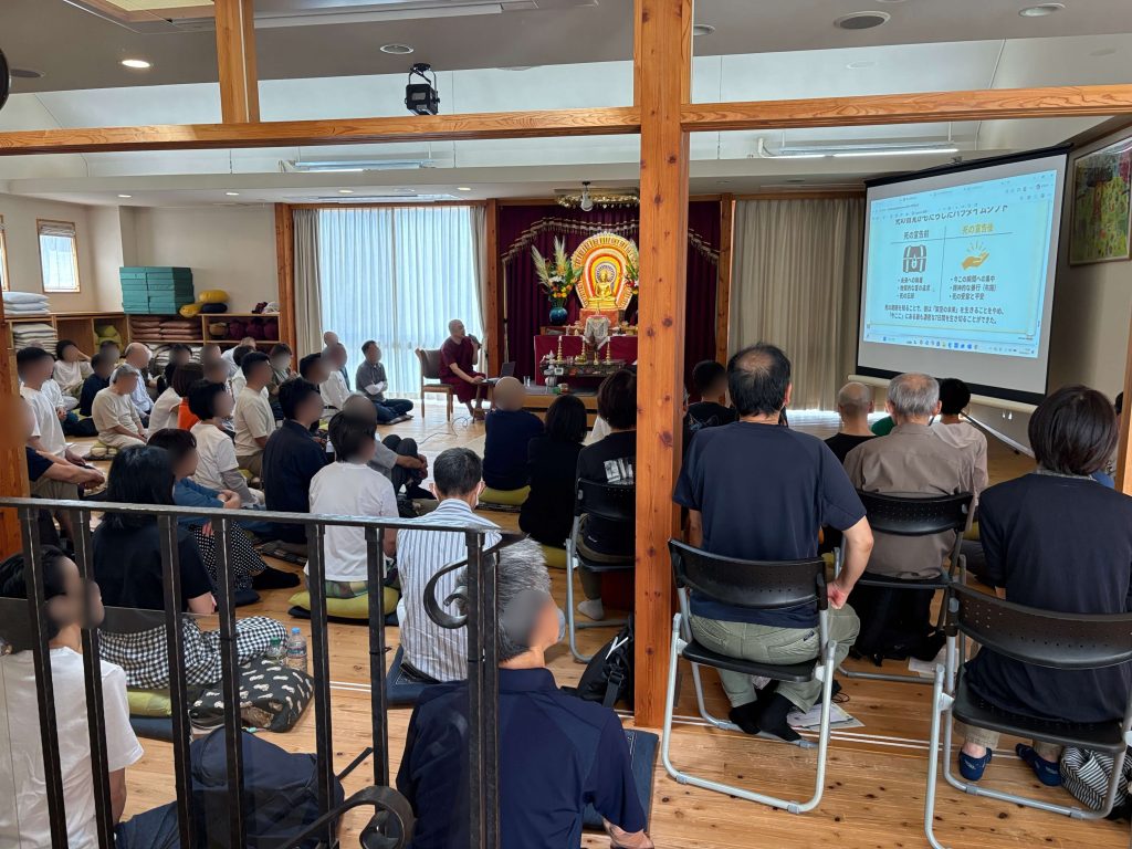 Buddhist ceremony in a temple with a monk speaking to a seated audience; a projector screen shows slides on the right.