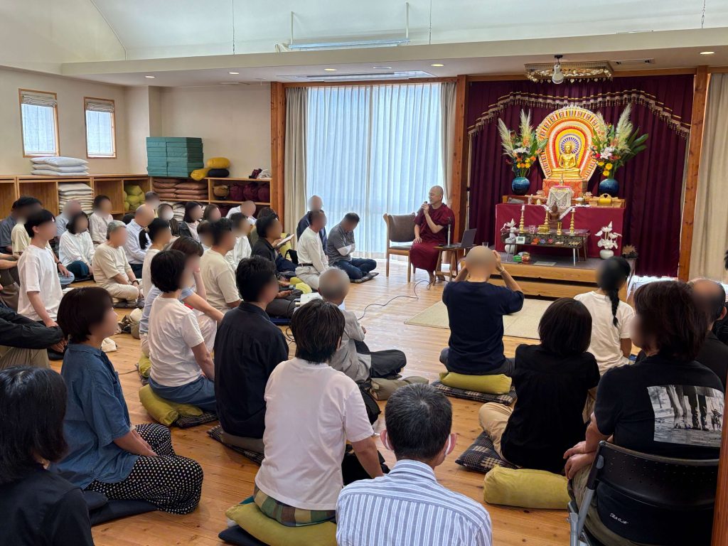 Monk in maroon robes speaks to a seated audience in a temple or meditation hall, people seated on cushions on a wooden floor.