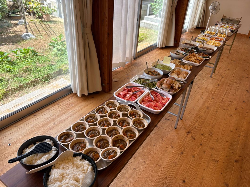 Buffet table with multiple white bowls and trays containing curry, rice, watermelon, bread, and salads in a bright room with wooden floors and sliding glass doors outside.