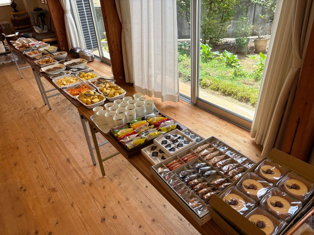 Buffet table with bowls of fruit, pastries, and snacks along a sunlit wooden floor by large glass doors opening to a garden.