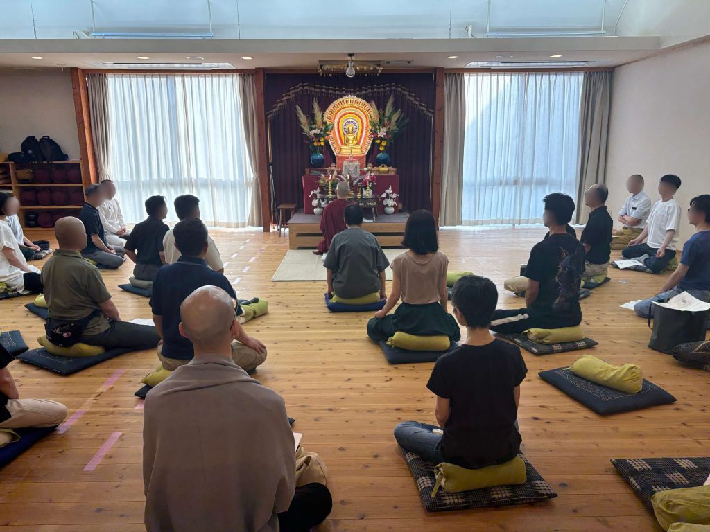 Group of people seated on cushions in a dojo-like room, facing a Buddha statue altar with flowers and offerings. A monk in maroon robes sits at the front center.