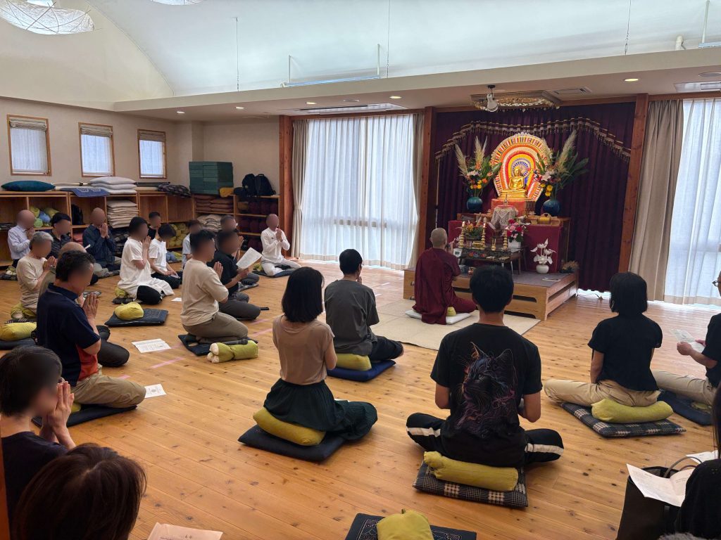 Group of people seated on cushions in a temple hall, meditating facing an ornate Buddha altar with flowers and drapes at the front.