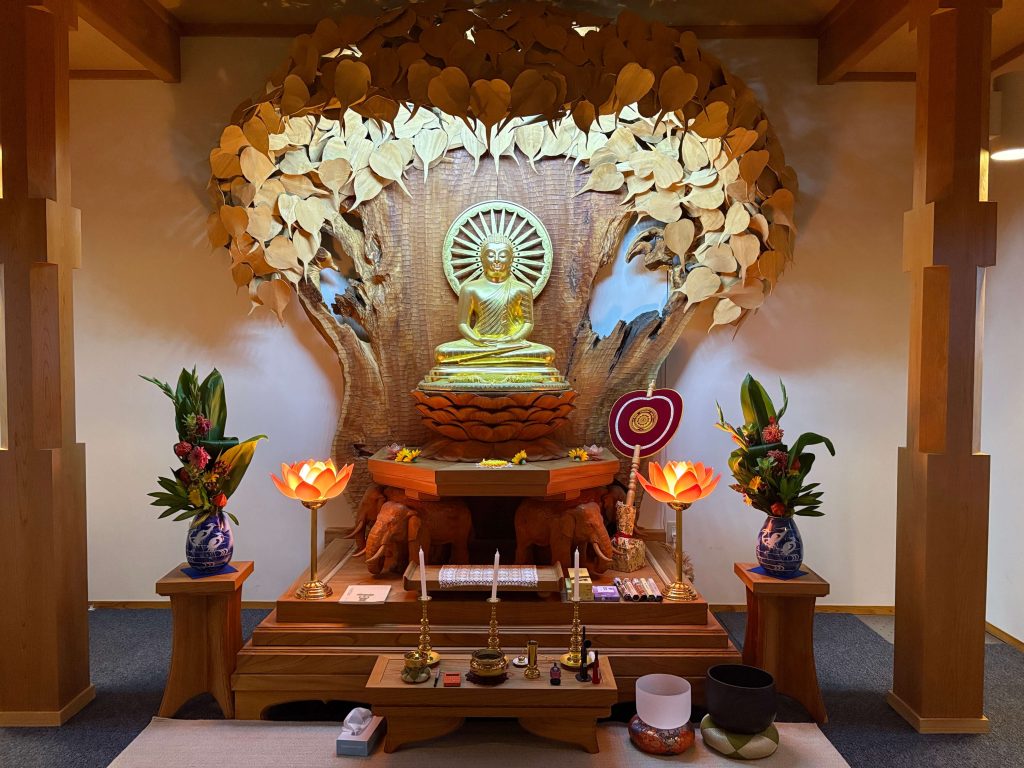 Golden Buddha statue seated on a multi-tiered altar, surrounded by a heart-shaped leaf canopy and lotus arrangements in blue vases.