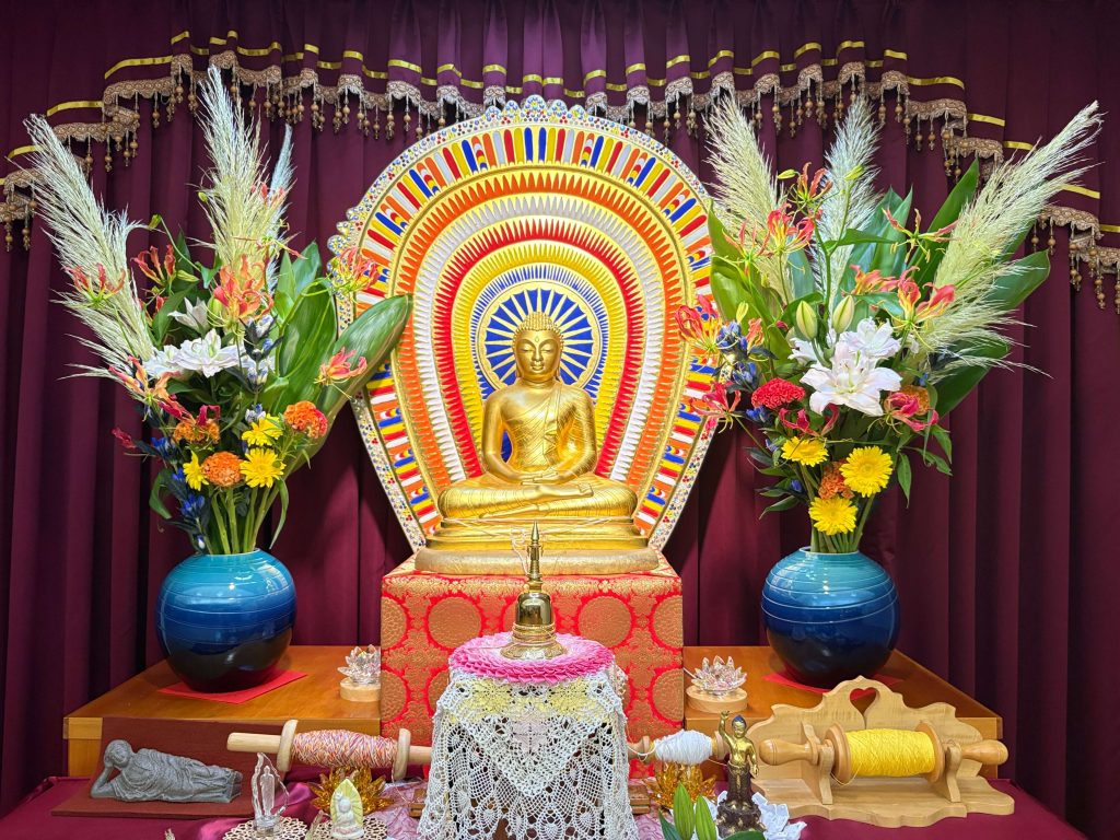 Golden Buddha statue seated in a colorful ceremonial mandala backdrop with flowers and decorative cloths on an altar